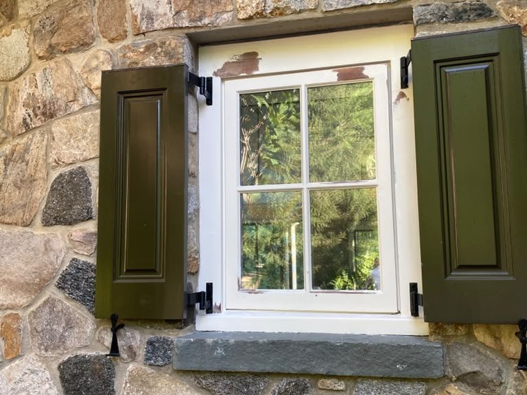 Wood window in a stone wall with green shutters, old paint peeling at the head before repainting