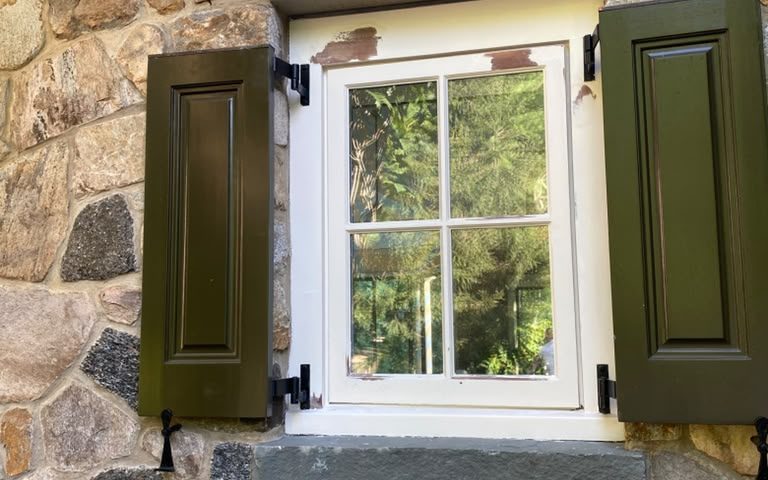 Wood window in a stone wall with green shutters, old paint peeling at the head before repainting