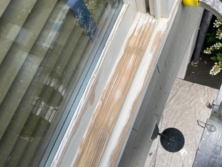 Exterior window sill partly scraped to bare wood, with masking tape on the glass