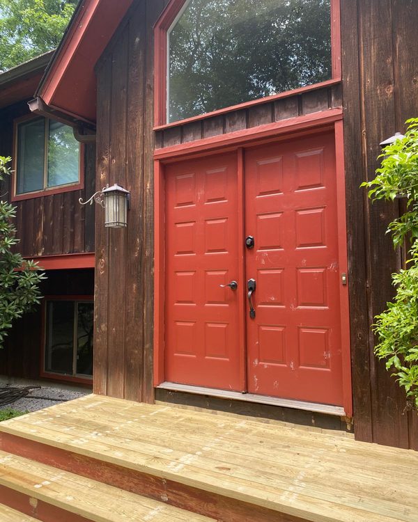 After: Red double front doors freshly painted, set in dark wood siding above a new deck