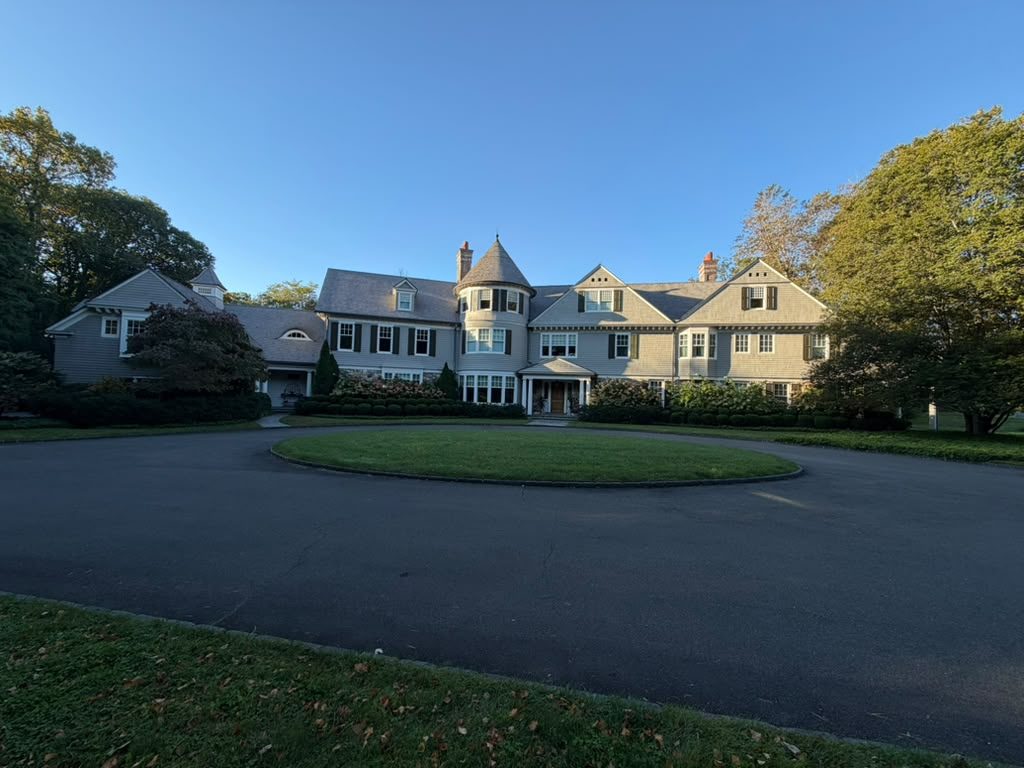 Large shingle-style home with a turret and dark shutters behind a circular drive