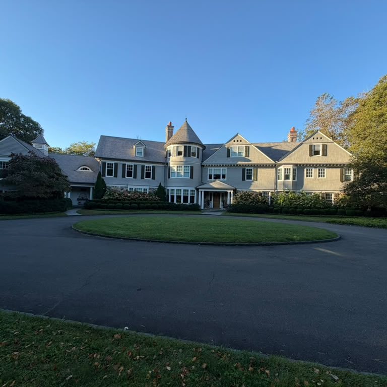 Large shingle-style home with a turret and dark shutters behind a circular drive
