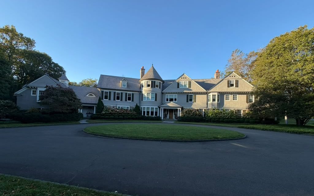 Large shingle-style home with a turret and dark shutters behind a circular drive